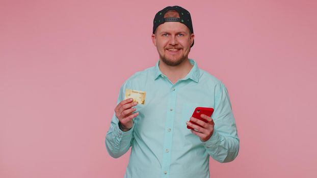 Sincere adult man customer using credit bank card and smartphone while transferring money, purchases online shopping, payment. Finance and internet. Young guy isolated alone on pink wall background