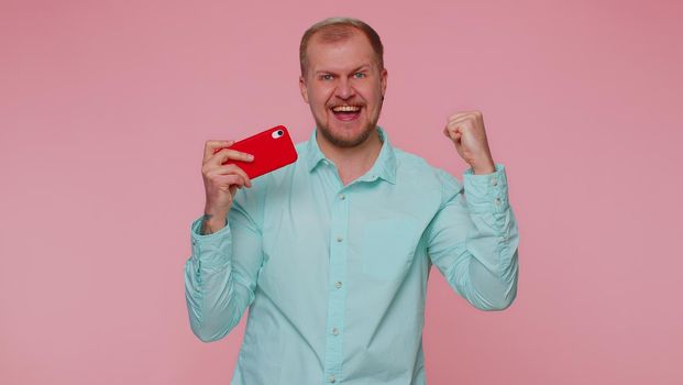 Worried funny addicted man in shirt enthusiastically playing racing video games on mobile phone. Young adult guy using smartphone gadget app with drive simulator isolated on pink studio background