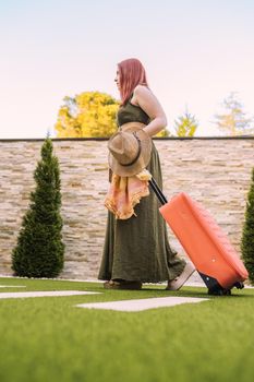 young woman traveller, entering hotel reception with her wheeled suitcase. woman on recreational trip. concept of rest and holiday. Outside, dim sunlight, grass floor with white tiles.