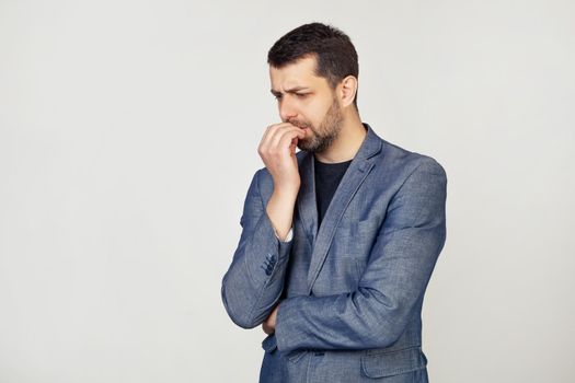 Young businessman with a smile, a man with a beard in a jacket, looks tense and nervous with hands on his lips, biting his nails. Anxiety problem. Portrait of a man on a gray background.