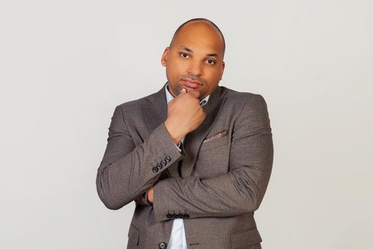 Young male African American businessman holding his chin with thoughtful facial expression looking into the camera. The guy is thinking about his future life. standing on a gray background