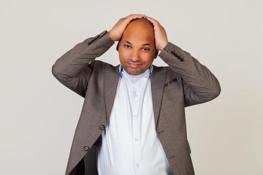 Portrait of a young African American guy businessman, holding his hands on his head, tired facial expression due to headache after a hard day at work. Standing on a gray background