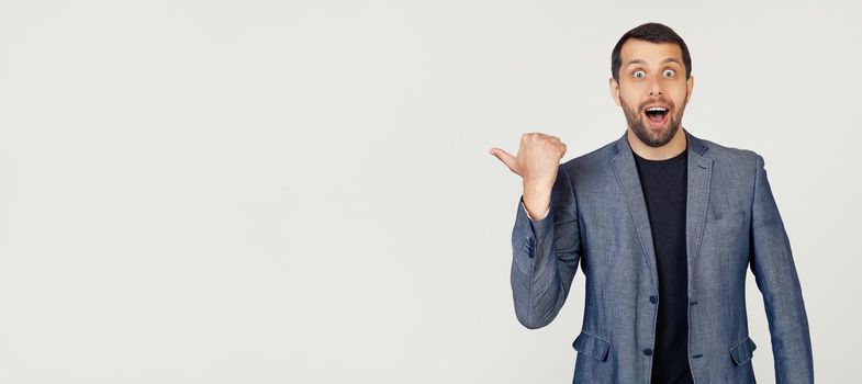 Young businessman man with a beard in a jacket, surprised, pointing finger to the side, with open mouth amazed expression. Portrait of a man on a gray background.