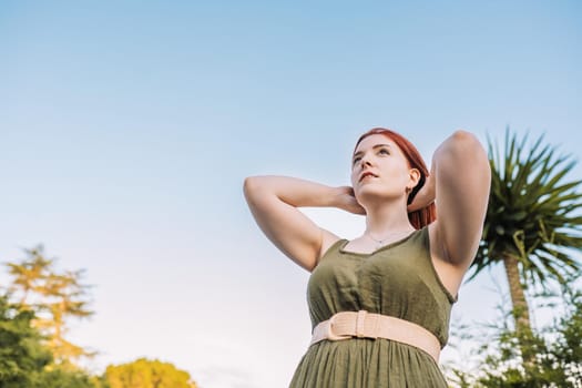 young adventurous girl, relaxing on her summer holiday. woman bathed in sunlight holding up her hair. concept trip and leisure. woman in green summer dress, straw hat, outdoors with natural light and plants in background.