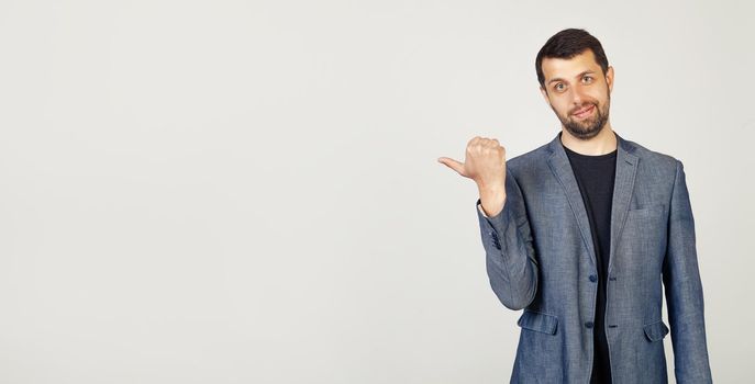 A young man with a beard smiling with a happy face, looking and pointing to the side with his thumb at a place for advertising, look here, on isolated background.