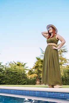 adventurous young woman, standing sunbathing in front of the swimming pool of her holiday hotel. woman on recreational trip. Outside, dim sunlight, grass floor with white tiles. vertical. copy space.