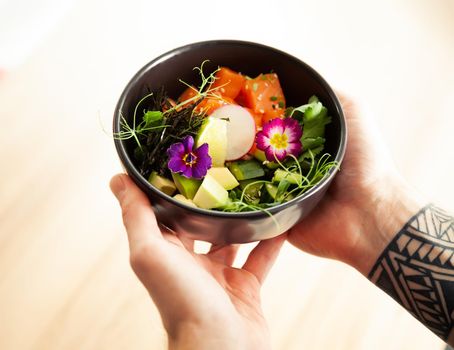 A man holds a bowl of Poke salad with salmon, avocado, cucumber, rice, Tamarind sauce, mango sauce, nori, lime, sesame, cilantro. Asian seafood salad concept
