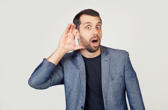 Young businessman man with a beard in a jacket, smiling with his hand above his ear, listening to rumors or gossip. Deafness concept. Portrait of a man on a gray background.