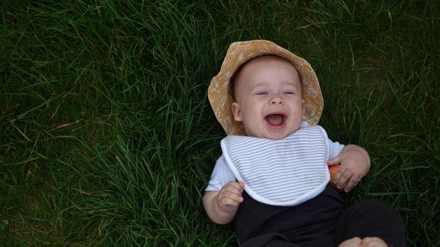 Small Happy Newborn child in summer panama hat Fall down laying on grass barefoot in Summer Sunny Day. Infant Kid Toddler Boy Smilling Face look at camera in Garden ouside Family Childhood Nature