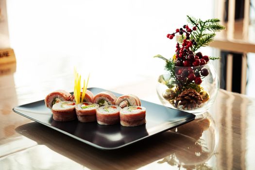 Sushi roll with bacon and fried sea bass on a black plate, ingredients fried sea bass, cream cheese, bacon, Iceberg salad, flying fish roe, Teriyaki sauce, rice, nori. For the restaurant menu