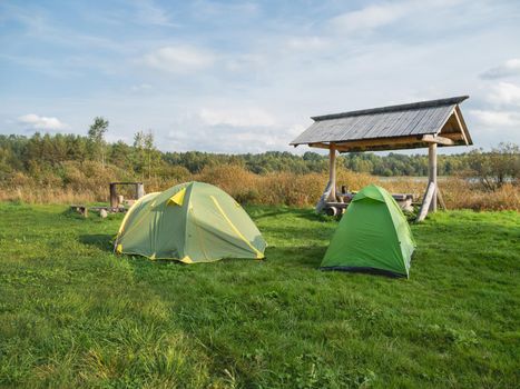 Two green touristic tents stand in camp. Wooden table, benches and fire pit for camping. Hiking in Russia.