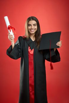 Front view of brunette girl graduating from college, university, high school. Pretty girl looking at camera, smiling, holding diploma, mortarboard. Isolated on red background,
