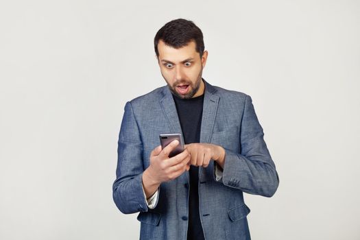 Young businessman man with a beard in a jacket using a smartphone scared shocked with a surprised face, scared and excited with an expression of fear. Portrait of a man on a gray background.