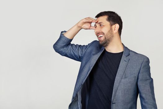 Young businessman man with a beard in a jacket, very happy and smiling, looking far away with hand above his head. Search concept. Portrait of a man on a gray background.