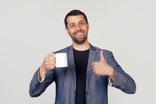 Young businessman with a smile, a man with a beard in a jacket, holding a cup of coffee and showing a thumb up gesture, a man with a happy face. Portrait of a man on a gray background.