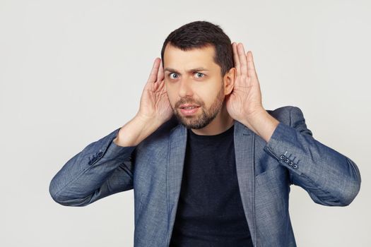 Young businessman man with a beard tries to hear both hands on his ear, curious about gossip. Hearing problems, deaf. Portrait of a man on a gray background.