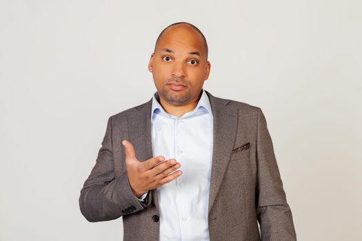 Young african american guy businessman looking at the camera with a surprised facial expression and gesturing with his hand. Standing on a gray background