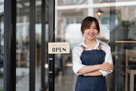 Beautiful asian young barista woman in apron holding tablet and standing in front of the door of cafe with open sign board. Business owner startup concept.