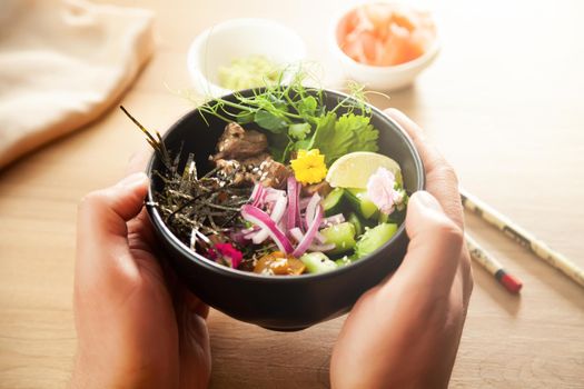 A man holds a Poke salad with beef in a bowl in his hands. Ingredients Beef, Nameko mushrooms, cherry tomatoes, rice, cucumber, red onion, sesame seeds, cilantro, lime. Asian salad concept.