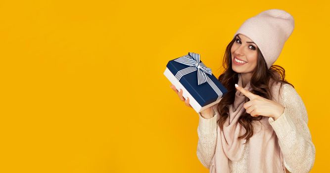 Happy young woman smiling pointing finger holding blue gift box in hand. A gift to your beloved man. A girl in a sweater looks at the camera and points to a gift. Christmas New Year holiday concept