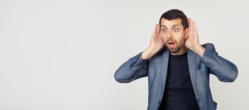 Young businessman man with a beard tries to hear both hands on his ear, curious about gossip. Hearing problems, deaf. Portrait of a man on a gray background.