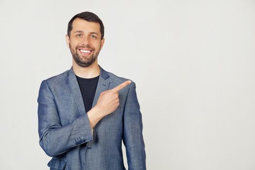 Young businessman man with a beard in a jacket, cheerful, with a smile on his face, pointing with his hand and finger to the side with a happy and natural expression. Portrait of a man on a gray background.