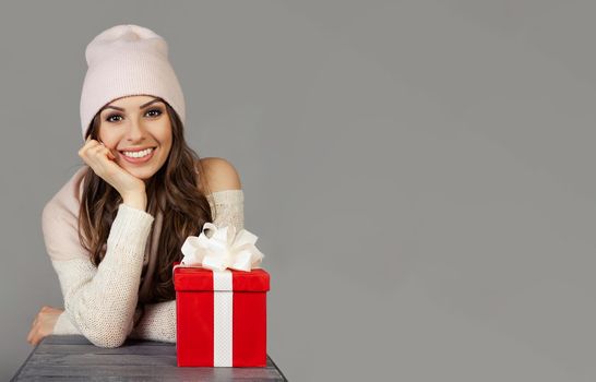 Smiling and happy, beautiful young woman in winter clothes with a red Christmas gift box on a gray background. The girl enjoys Christmas shopping, meets the New Year. Shopping sale concept.