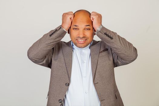 Unhappy male african american businessman in a jacket holding his hands on his head being very disappointed and looking at the camera upset because of the bad news, on a gray background.