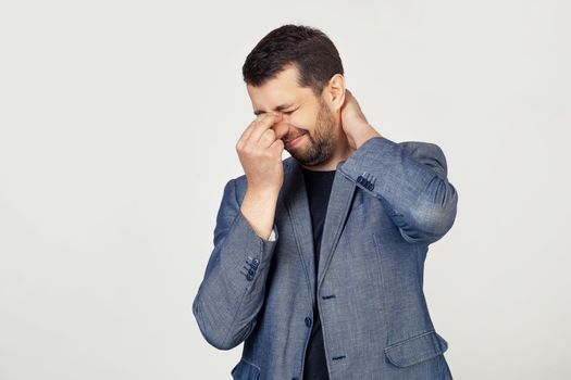 Young businessman man with a beard in a jacket, tired of rubbing his nose and neck, feeling tired and headache. Stress and frustration concept. Portrait of a man on a gray background.