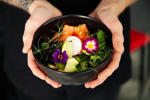A man holds a bowl of Poke salad with salmon, avocado, cucumber, rice, Tamarind sauce, mango sauce, nori, lime, sesame, cilantro. Asian seafood salad concept