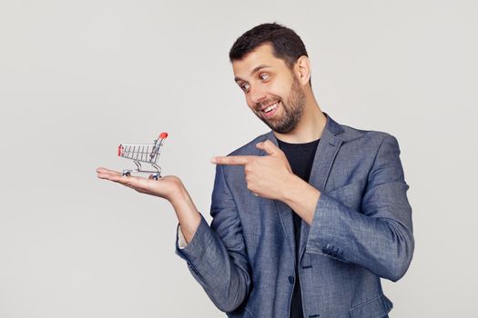 Young businessman man with a beard in a jacket looks and points to a shopping cart on his hand, showing with his hand and pointing with his finger. Portrait of a man on a gray background.