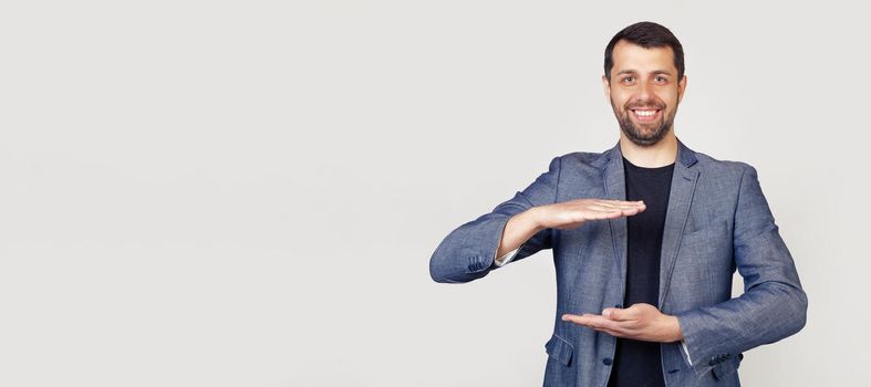 Young businessman man with a beard in a jacket, gesturing with his hands showing a large and large size sign, a symbol of measure Smiling looking into the camera Portrait of a man on a gray background