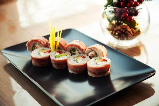 Sushi roll with bacon and fried sea bass on a black plate, ingredients fried sea bass, cream cheese, bacon, Iceberg salad, flying fish roe, Teriyaki sauce, rice, nori. For the restaurant menu
