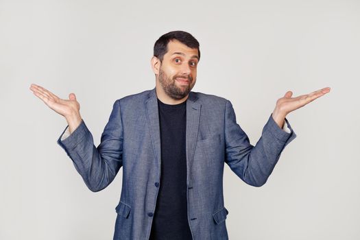 A young businessman man with a beard in a jacket, shrugs his shoulders, arms outstretched complete disbelief, doubts to say the answer I have no idea I dont know Portrait of a man on a gray background