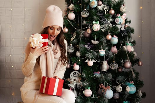 Smiling and happy, beautiful young woman in winter clothes with a red Christmas gift box on the background of a decorated Christmas tree. The girl delightedly opens the gift, greets the New Year.