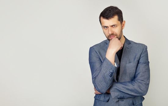 The man is looking thoughtfully at the camera. Confident young man with a beard in a stylish jacket, holds his hand on his chin, raised eyebrow thinks about something makes a plan, ponders a cool idea
