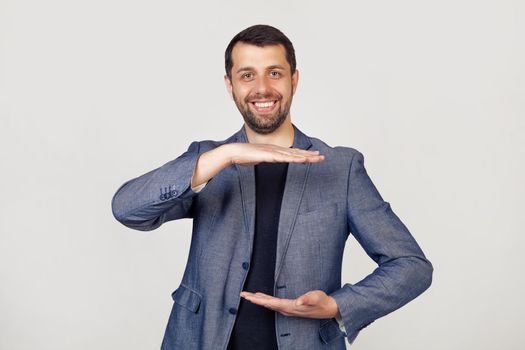 Young businessman man with a beard in a jacket, gesturing with his hands showing a large and large size sign, a symbol of measure Smiling looking into the camera Portrait of a man on a gray background