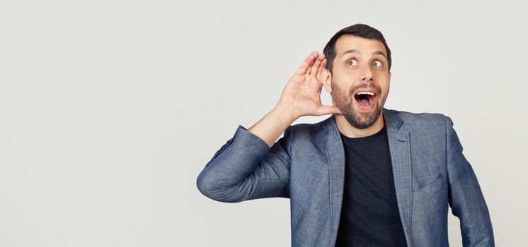 Young businessman man with a beard in a jacket, smiling with his hand above his ear, listening to rumors or gossip. Deafness concept. Portrait of a man on a gray background.