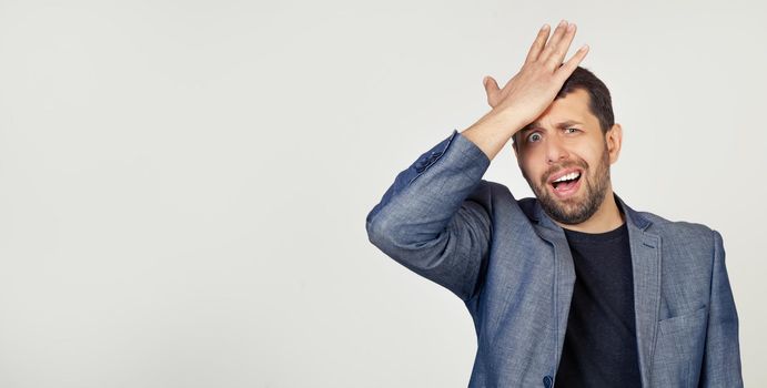 Young businessman man with a beard in a jacket, panics because of a forgotten deadline, putting his hand on his head for a mistake feels stress, forced to cover up the mess or mistake. gray background