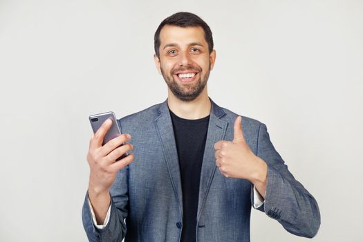 Young businessman with a smile, a man with a beard in a jacket, with a smartphone in his hand, happy with a big smile, makes an OK sign, thumb up with fingers, a great sign.