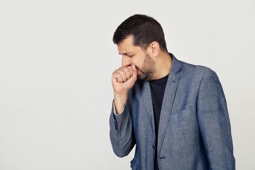 Young businessman man with a beard in a jacket, feeling unwell and coughing as a symptom of a cold or bronchitis. Health care concept. Portrait of a man on a gray background.
