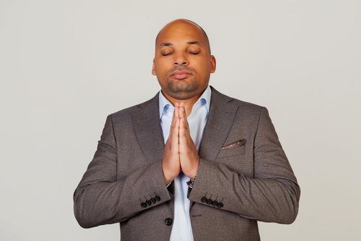 Handsome young concentrated african american businessman male in jacket holding hands together in front of face, eyes closed praying and hoping today is a good day, on gray background
