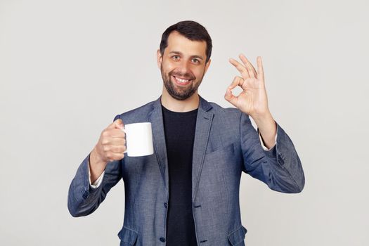 Young businessman with a smile, a man with a beard in a jacket, holding a cup of coffee and showing the gesture everything is ok, a man with a happy face. Portrait of a man on a gray background.