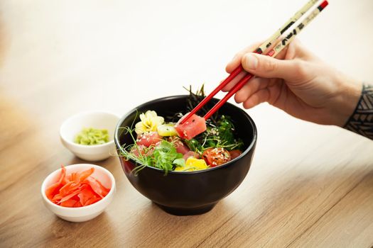 A man eats a Poke salad with tuna in a bowl with chopsticks. Place the tuna salad in a bowl. Young man guy in a restaurant eating poke salad. Asian seafood salad concept