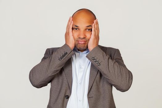 Young african american guy businessman holding his head in hands with unhappy facial expression looking sad and trying not to cry. Standing on a gray background