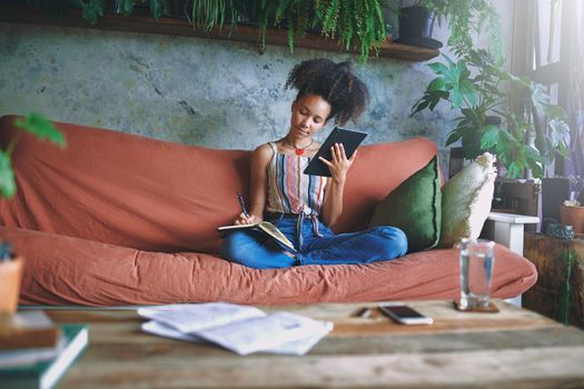 Beautiful African woman scrolling on her tablet and making notes in her living room