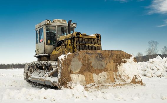 Yellow crawler tractor bulldozer with a bucket works in winter clearing the road from snow. Frosty day with bright sun and beautiful blue sky in the background. Yellow bulldozer in winter