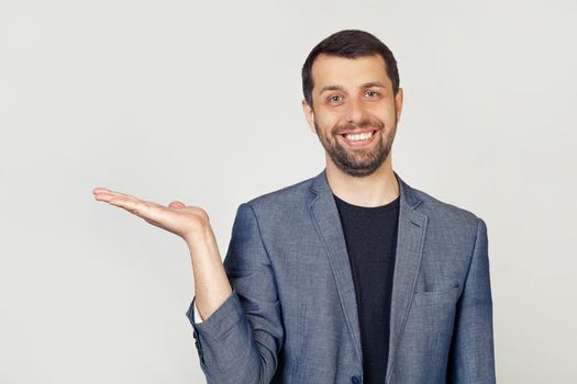 Young man with a beard in a white t-shirt smiling, cheerful presenting and pointing with his palm, looking at the camera. Stands on isolated yellow background.