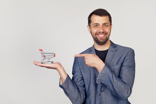 Young businessman man with a beard in a jacket looks at the camera and points to the shopping cart on his hand, showing with his hand and pointing with his finger