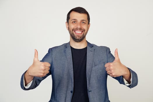 Young businessman man with a beard in a jacket, a sign of success makes a positive gesture with his hand, thumbs up smiling and happy. Cheerful expression and winner gesture. gray background